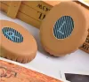 Close-up of two brown ear pads with a blue and black striped pattern inside the oval opening. A wooden crate with text is in the background. Produktbild-Vorschau