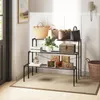 A three-tiered black metal shelf unit displaying various items including handbags, decorative boxes, and a plant, placed on a rug in a room with white paneled walls. Podgląd zdjęcia produktu