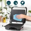 A hand is cleaning a panini grill with a blue cloth. The grill is open, showing the ribbed cooking plates. In the background, there are plants and a bottle of oil. Produktbild-Vorschau