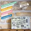 A flat, rectangular tin box with a vintage-style illustration of various items, placed on a wooden surface against a brick wall background. Text overlays describe the product's features. Voir les images du produit