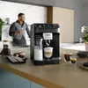 A man is in the background of a kitchen scene, holding a drink, while a coffee machine is in the foreground. Produktbild-Vorschau
