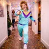 A man in a retro tracksuit is pointing at the camera in a hallway decorated with confetti and streamers. Produktbild-Vorschau