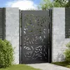 A black metal garden gate with a decorative floral pattern, set between two brick pillars and a wooden fence. Náhľad obrázku produktu