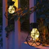 Two solar-powered garden lanterns with a diamond-shaped metal cage and a light bulb inside, hanging near a fence and plants at dusk. Produktbild-Vorschau