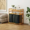 A wooden laundry cart with three black fabric bins and two shelves, placed in a room with a sofa and a plant. Anteprima dell’immagine prodotto