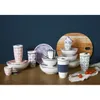 A collection of ceramic tableware and travel mugs with geometric patterns, arranged on a wooden surface against a dark blue background. Produktbild-Vorschau