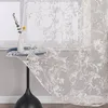 A sheer curtain with a floral pattern is draped over a table with a vase and a single flower. Produktbild-Vorschau