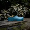 A teal clog sits on a weathered wooden surface with lush green foliage in the background. Produktbild-Vorschau