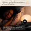 A woman is lying in bed, looking towards a Lumie light alarm clock that is emitting a warm, sunrise-like glow. Produktbild-Vorschau