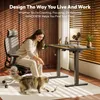 A woman is sitting in an office chair, playing with a cat in front of a standing desk. Produktbild-Vorschau
