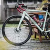 A bicycle parked outdoors on a wet surface, leaning against a wooden table. Produktbild-Vorschau