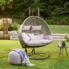 A hanging egg chair with cushions and a throw blanket sits on a grassy lawn in a garden setting, with stairs and plants in the background. Náhled obrázku produktu