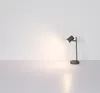 A modern desk lamp with a gray metal finish is placed on a light gray surface in front of a white wall. Produktbild-Vorschau