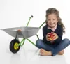 A young girl is sitting next to a toy wheelbarrow, holding an apple. Produktbild-Vorschau