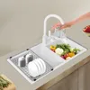 A person's hand is shown rinsing vegetables in a modern white double sink with a white faucet. A dish rack with plates and bowls is to the left of the sink. Podgląd zdjęcia produktu