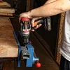 A person is using a cordless drill to drill into a metal bar clamped in a vise on a wooden workbench. Produktbild-Vorschau