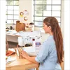 A woman is using a sewing machine on a wooden table, with shelves of craft supplies in the background. Produktbild-Vorschau