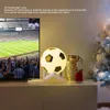 A soccer ball lamp is displayed on a shelf next to a television showing a soccer game, with decorative items on the right. Produktbild-Vorschau