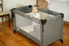 A baby is standing in a gray travel crib in a room with wooden floors. Anteprima dell’immagine prodotto