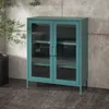 A teal metal display cabinet with glass doors stands on a dark floor in front of a concrete wall. Produktbild-Vorschau