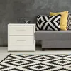 A white nightstand with two drawers next to a gray sofa with decorative pillows and a black and white geometric rug in front. Náhled obrázku produktu