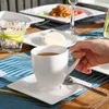 A person's hand is holding a white coffee cup filled with coffee, placed on a saucer. The setting appears to be an outdoor dining area with plates of food and a glass of water visible in the background. Produktbild-Vorschau