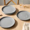 Four gray plates are arranged on a wooden table, with two clear glasses and a folded white cloth also visible. Produktbild-Vorschau