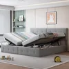 A gray upholstered bed frame with the mattress lifted, revealing storage space underneath. The bed has a tufted headboard and is positioned in a room with blinds, shelves, and artwork. Náhľad obrázku produktu