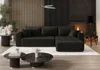 A dark gray sectional sofa with tufted cushions is placed on a white rug in a modern living room setting, with a fireplace and modern decor. Produktbild-Vorschau
