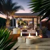 Four LED tea lights are placed on a wooden surface in the foreground, with a blurred background of a modern outdoor living space at dusk. Produktbild-Vorschau