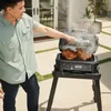 A man is using a black electric grill outdoors, with two chickens cooking on it. Voir les images du produit