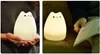 A collage of two images showing a white cat-shaped LED night light. The left image shows hands holding the lamp, and the right image shows the lamp on a couch next to a person reading. Produktbild-Vorschau