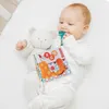 A baby lying on a white surface, playing with a soft toy and a fabric book. Anteprima dell’immagine prodotto
