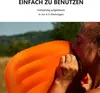 A person is shown inflating an orange camping pillow outdoors. Produktbild-Vorschau