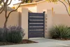 A modern gray aluminum garden gate with horizontal slats, set within a beige stucco wall and flanked by plants and a tree. The gate is in a residential outdoor setting. Produktbild-Vorschau