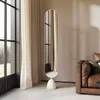 A modern oval floor mirror with a beige base stands next to a large window with sheer curtains, and a brown leather sofa is visible on the right. Produktbild-Vorschau