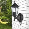 A black outdoor lantern with a motion sensor is mounted on a white brick wall, with a garden and greenery in the background. Produktbild-Vorschau