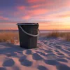 A dark gray bucket sits on a sandy beach at sunset, with tall grass and the ocean in the background. Anteprima dell’immagine prodotto