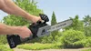 Hands holding a cordless chainsaw in an outdoor setting with trees and bushes in the background. Náhľad obrázku produktu