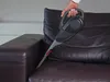 A hand is using a handheld vacuum cleaner to clean a brown leather sofa. Anteprima dell’immagine prodotto