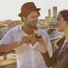 A man and a woman toasting with drinks on a rooftop with a city skyline in the background. Produktbild-Vorschau