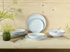 A set of porcelain dinnerware is arranged on a wooden table, with a vase of greenery in the background. Produktbild-Vorschau