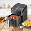 A black digital air fryer is shown on a wooden table, with a basket full of chicken wings pulled out. To the left, a plate of steak and fries is visible, and to the right, a plate of fries and a small bowl of ketchup are present. The background shows a blu Produktbild-Vorschau