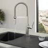 A modern kitchen faucet with a flexible spout and pull-down spray head is shown in use, with water flowing into a dark sink. The background features a large window with a city view and a potted plant. Anteprima dell’immagine prodotto