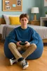 A young person sits on a large blue beanbag chair in a bedroom, holding a video game controller. Voir les images du produit