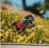 A red and black remote-controlled car is captured mid-air, appearing to jump over a ramp or obstacle, with a blurred background of yellow flowers and green foliage. Podgląd zdjęcia produktu