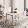 A white dining table set with four chairs, set on a patterned rug in a well-lit room. Produktbild-Vorschau