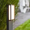 A modern outdoor LED path light is shown next to lush green foliage, with a blurred pathway and outdoor furniture in the background. Anteprima dell’immagine prodotto