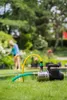 A garden pump is set up on a lawn with hoses connected, while a person waters plants in the background. Produktbild-Vorschau