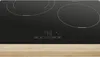 A close-up shot of a black induction cooktop with white heating elements and control buttons, resting on a light wooden surface. Voir les images du produit